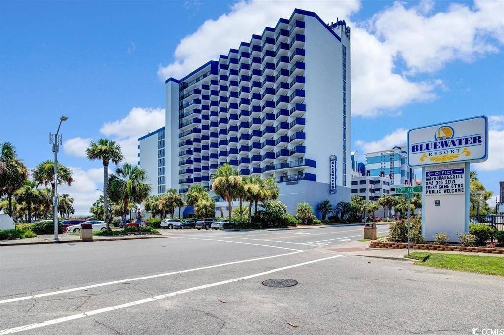 A tall, blue and white beachfront hotel; palm trees line the road and sign in the foreground.