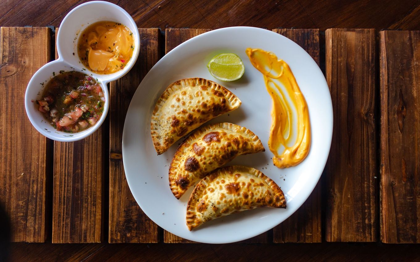 Empanadas with sauces and lime on a white plate, set on a wooden table.