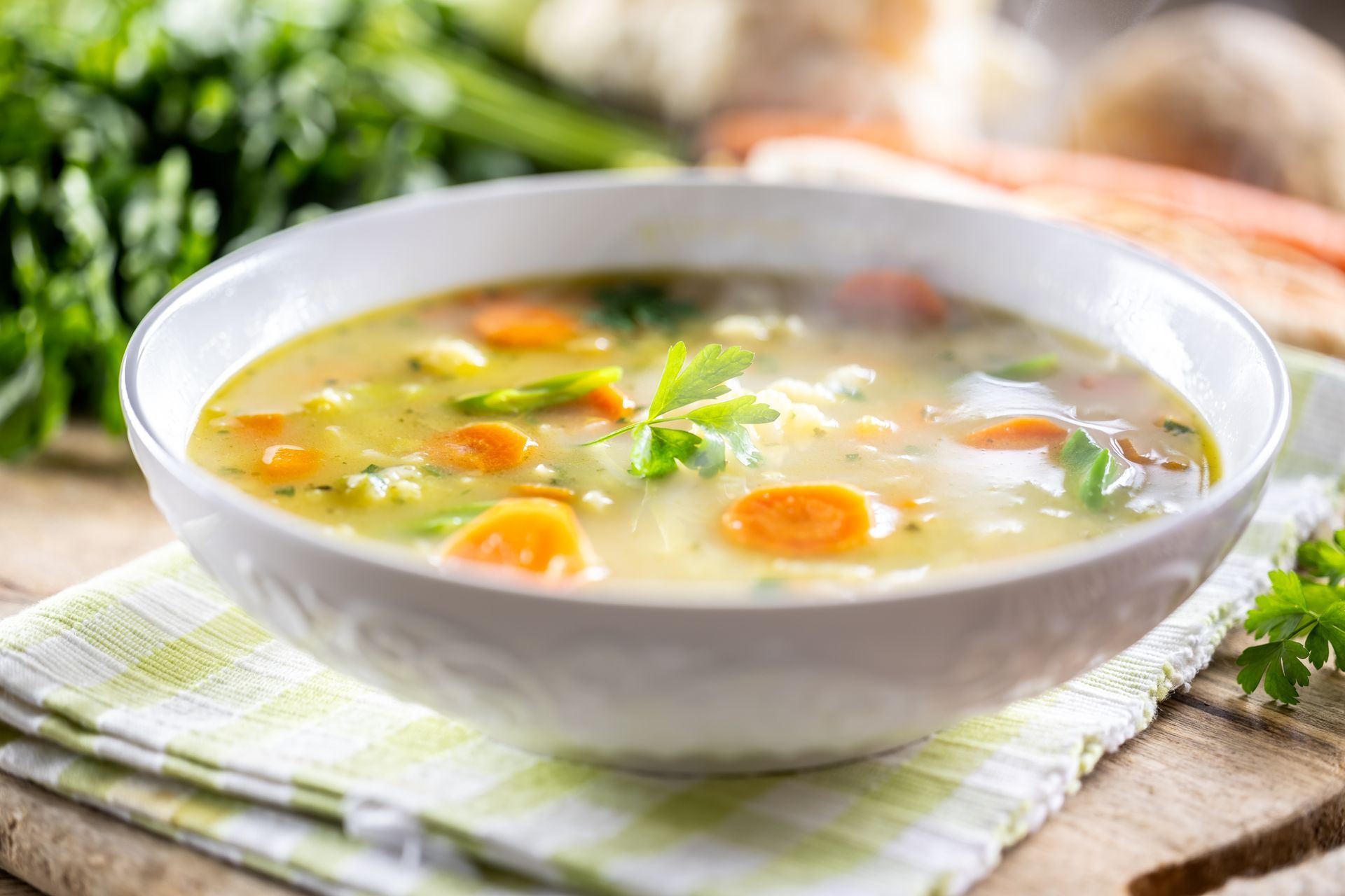 Chicken noodle soup in a white pot with ladle, vegetables, herbs, and a side of spices.