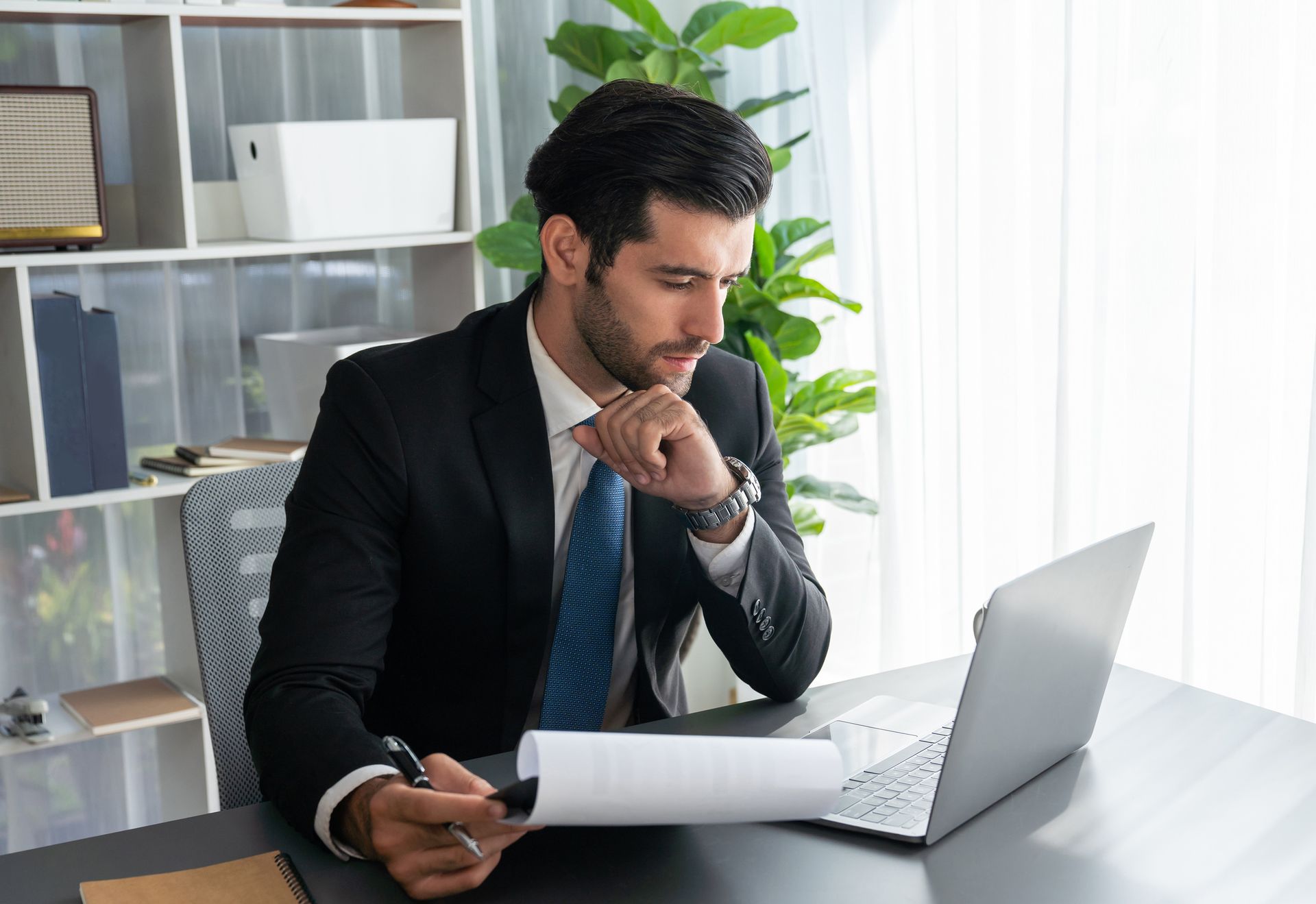 A man in a suit and tie is sitting at a desk in front of a laptop computer.