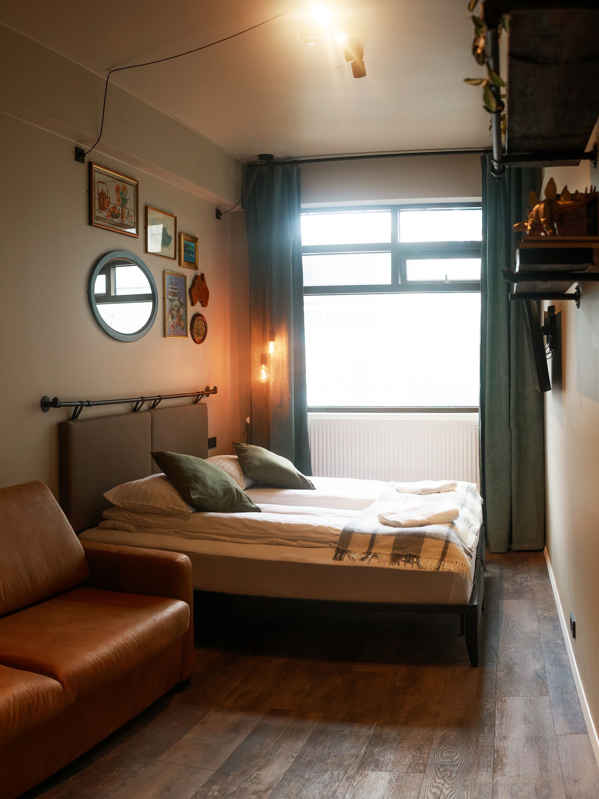 Small bedroom with bed, sofa, art on the wall, and a large window with green curtains.