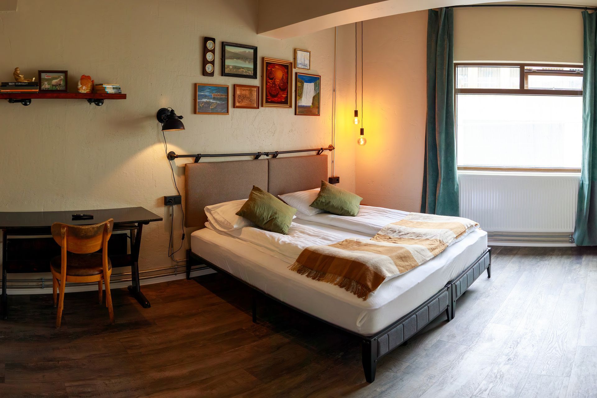 Cozy bedroom with bed, desk, and artwork on the wall; teal curtains and a window on the right.