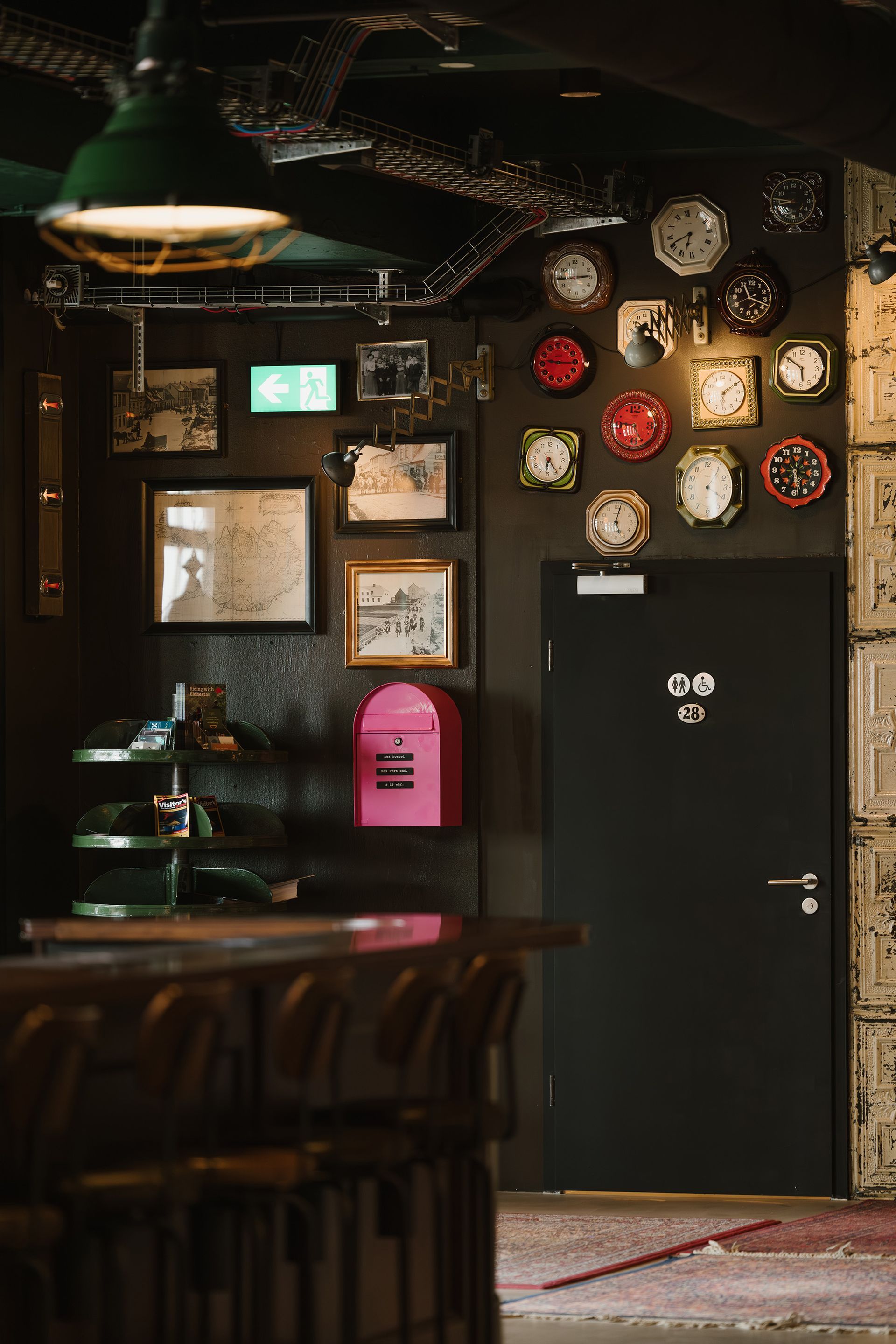 Dark interior with wall of clocks, framed pictures, and pink mailbox next to a black door.