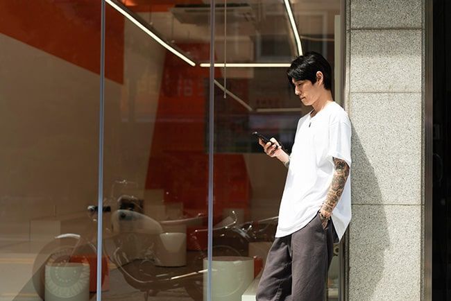 Un hombre con camisa blanca se apoya en un edificio, mirando el teléfono. Se ven tatuajes en el brazo.
