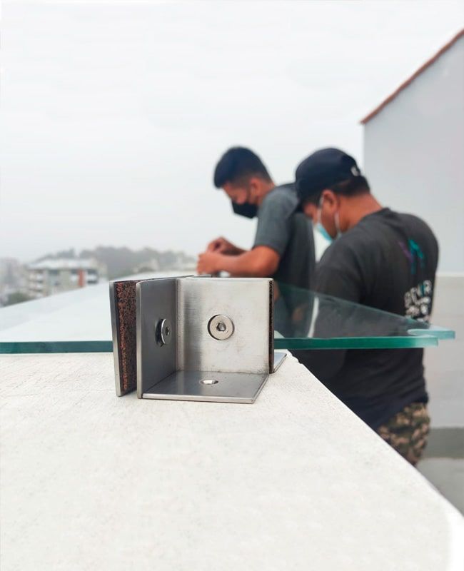 Dos trabajadores instalando paneles de vidrio en el borde de un edificio; soporte de acero inoxidable en primer plano.