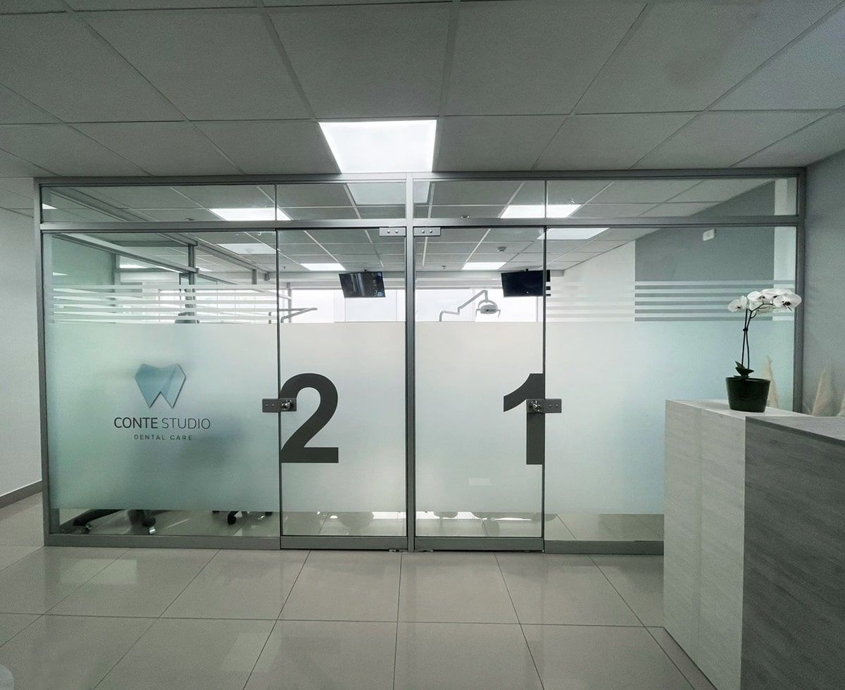 Entrada al consultorio dental con puertas de vidrio, ventanas esmeriladas y números 1 y 2. Paredes blancas y decoración moderna.