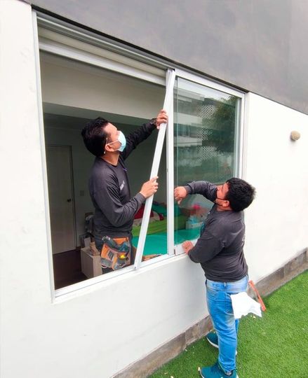 Dos personas instalando una mosquitera blanca en una ventana corrediza de vidrio.
