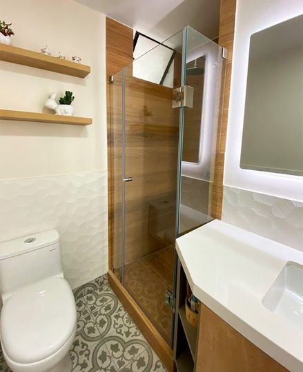 Baño con inodoro, ducha, detalles de madera y estanterías decorativas. Suelo de baldosas blancas y grises.