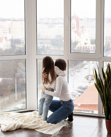 Mujer arrodillada abrazando a un niño junto a una gran ventana con vistas al paisaje urbano.