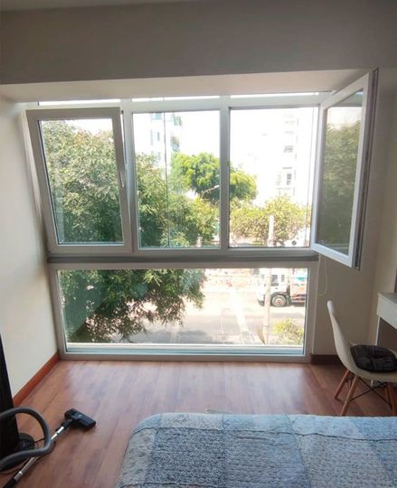 Recámara con amplios ventanales con vista a la calle y árboles, parcialmente abierto; piso de madera, paredes blancas y una silla.