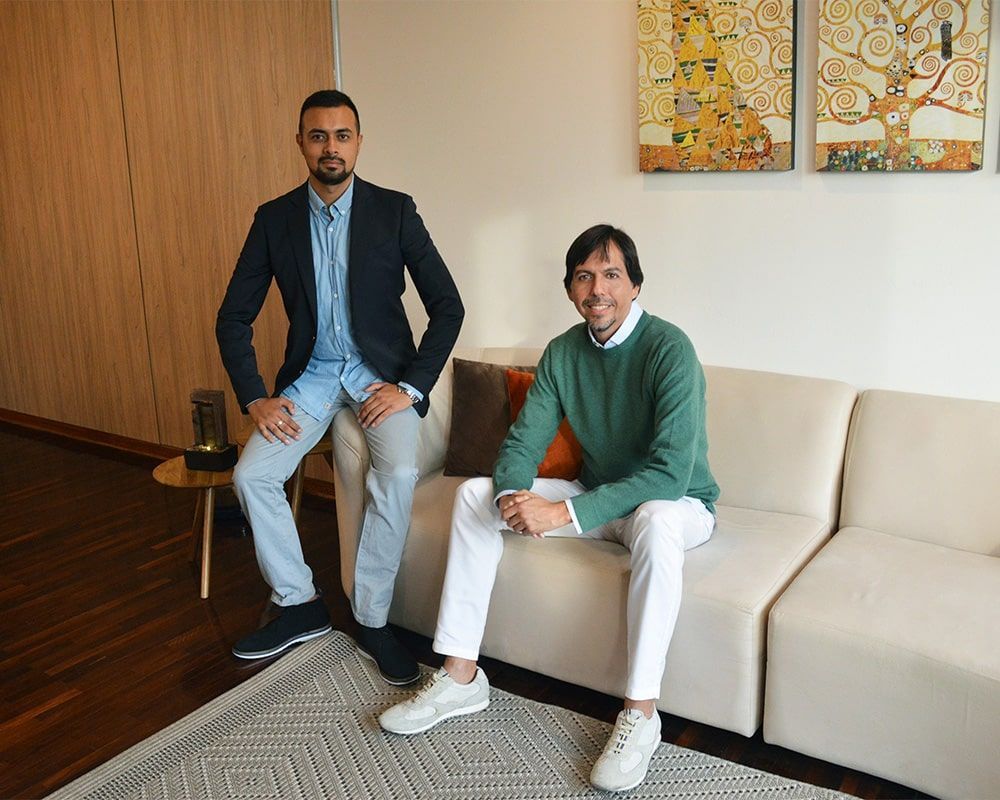 Dos hombres sentados en un sofá posando para una foto. Uno lleva un blazer, el otro, un suéter verde. Habitación en tonos neutros con obras de arte.