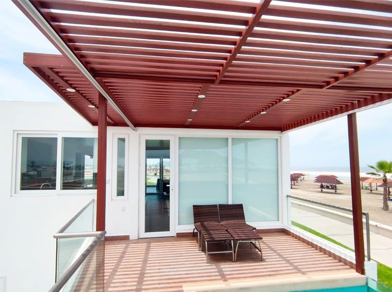 Deck con pérgola roja, dos sillas tipo lounge y vista parcial a la piscina y al océano.