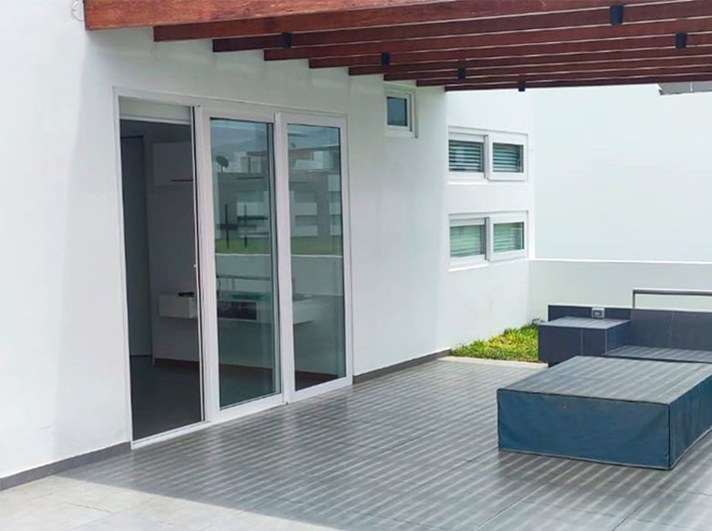 Patio con puertas corredizas de vidrio, paredes blancas y una pérgola de madera oscura. Los muebles de exterior están cubiertos.