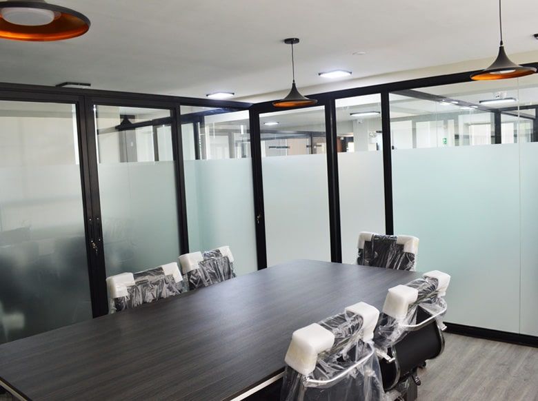 Sala de reuniones con mesa grande oscura, sillas, paredes de vidrio esmerilado, molduras negras y luces colgantes.