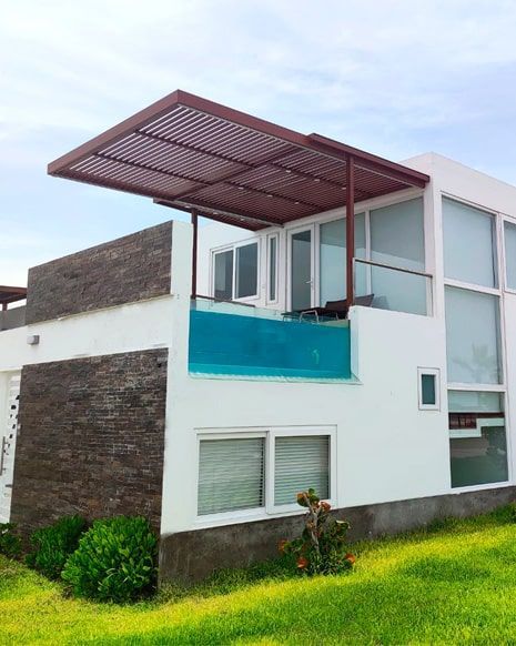 Casa blanca con pared de piedra marrón y pérgola, barandilla de balcón azul, con vista al césped verde.