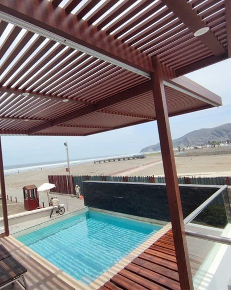 Piscina y patio con vista a la playa, pérgola marrón, terraza de madera, agua azul, playa de arena, cielo nublado.