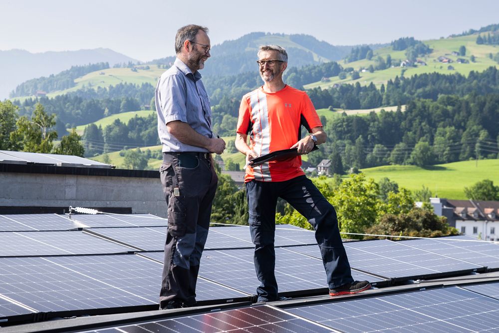 Zwei Männer auf einem Dach mit Solarmodulen und Bergen im Hintergrund besprechen Pläne.