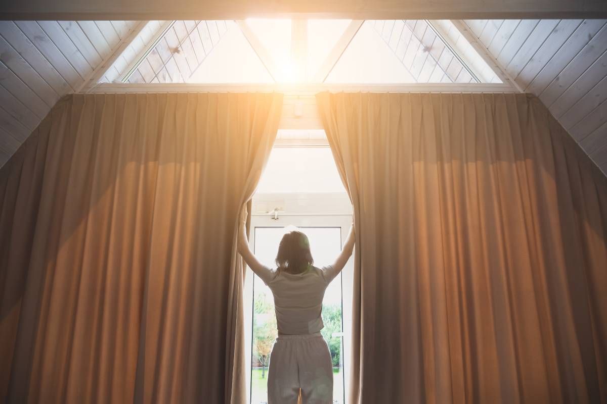A person opening earthy-toned interior fabric drapes to let sunshine into their modern home at Curtain Concepts near Huntington, WV