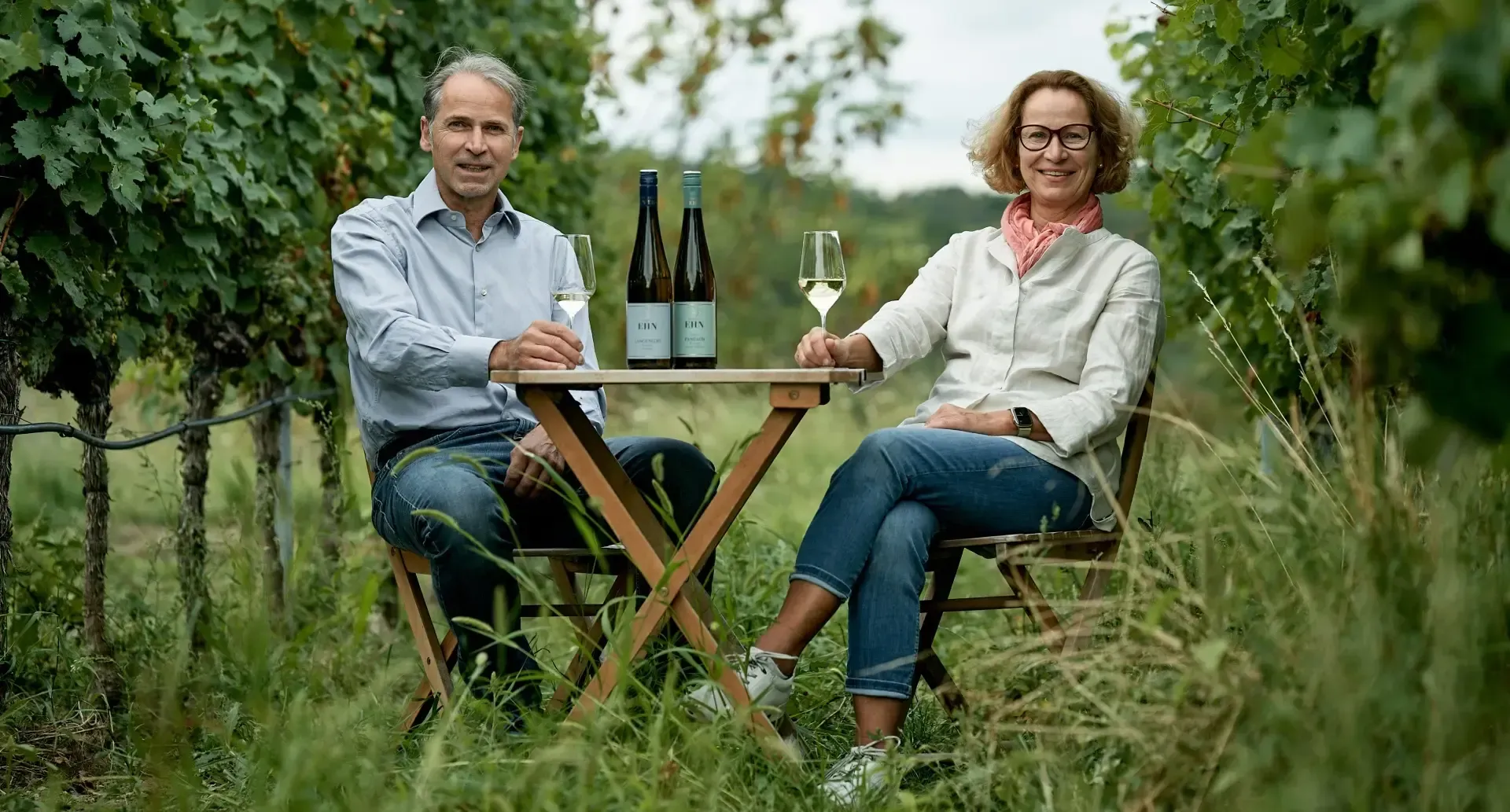 Ein lächelndes Winzerpaar Ehn sitzt an einem kleinen Klapptisch aus Holz mitten in einer grünen Rebenreihe. Auf dem Tisch stehen zwei Weinflaschen des Weinguts Ludwig Ehn, während beide jeweils ein Glas Weißwein in der Hand halten.