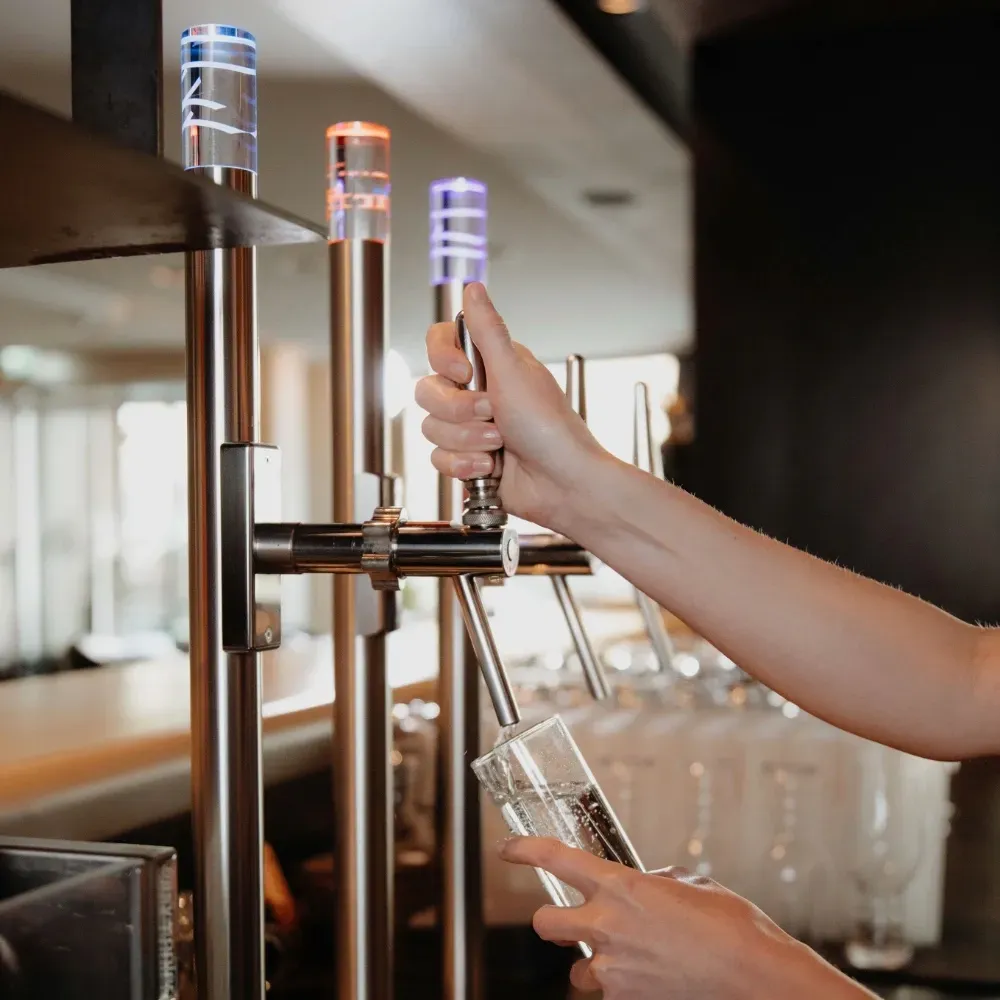 Eine Nahaufnahme zeigt eine Hand, die einen Hebel einer professionellen, metallenen Wellwasser-Zapfanlage in einem Restaurant bedient. Der Zapfhahn füllt ein darunter gehaltenes, klares Wasserglas mit prickelndem Wasser. Oben an der Anlage befinden sich drei säulenförmige Auslässe mit leuchtenden Lichtelementen in den Farben Blau, Rot und Violett. Der Hintergrund ist unscharf und lässt ein modernes Restaurantambiente mit Tischen und einer Fensterfront erahnen. Die Szene ist gut beleuchtet und fokussiert auf den Zapfvorgang.