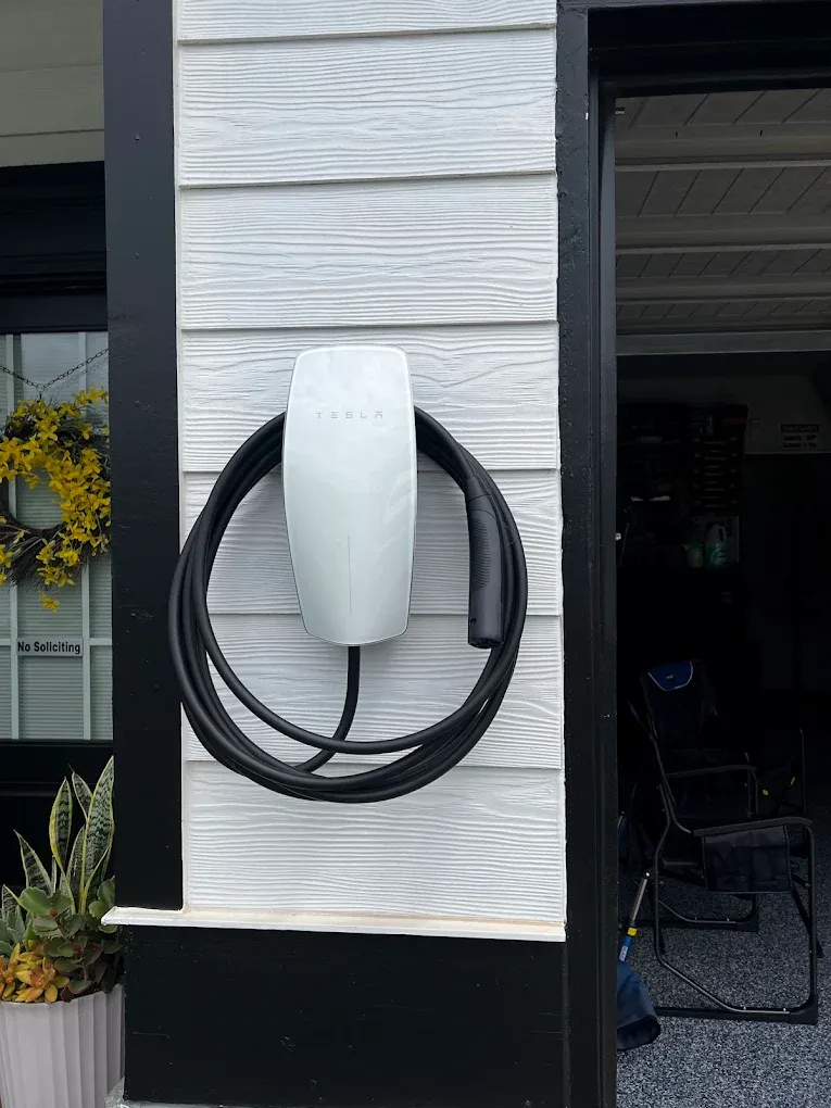Wall-mounted white EV charger with black cable coiled beside a house exterior