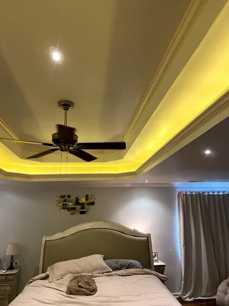 Bedroom with a lit tray ceiling, ceiling fan, gray walls, and a large bed with beige headboard and curtains.