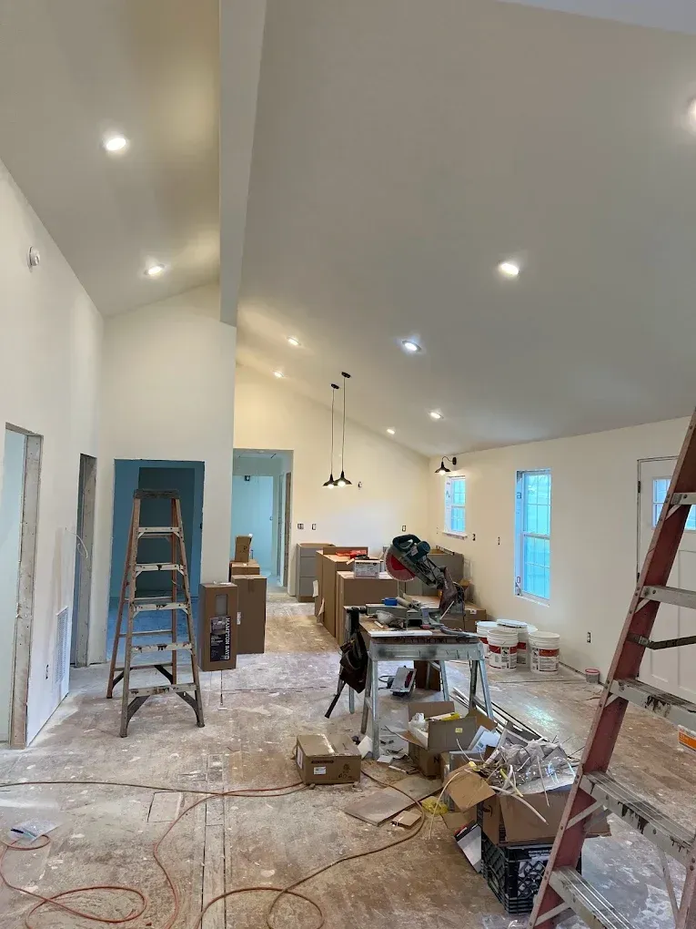 Renovation site in a bright room with ladders, tools, paint supplies, and unfinished flooring.
