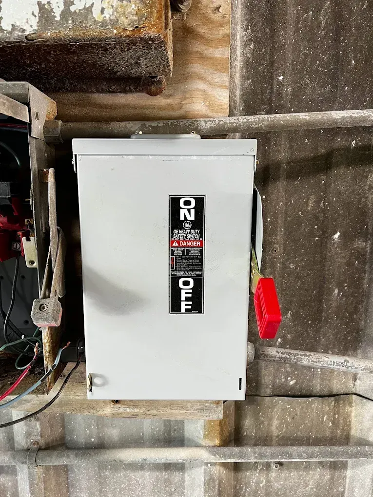 Gray electrical panel mounted on a concrete wall in a utility area, with a red handle on the right.