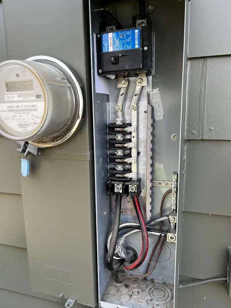 Open electrical meter panel with breakers and wiring exposed beside a gray utility meter.