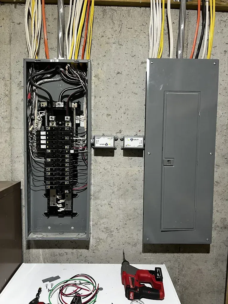 Open electrical panel next to a closed gray breaker box, with tools and wires on a work table.
