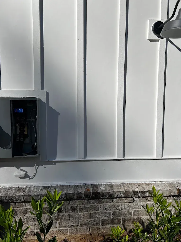 Outdoor wall with black utility box, silver siding, brick base, and shrubs in front.