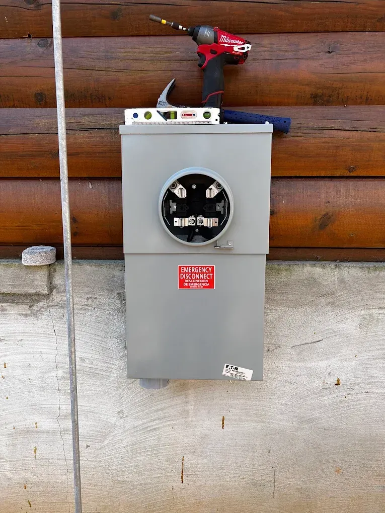 Gray electrical meter box with round dial, mounted on a concrete wall under a wooden fence board.