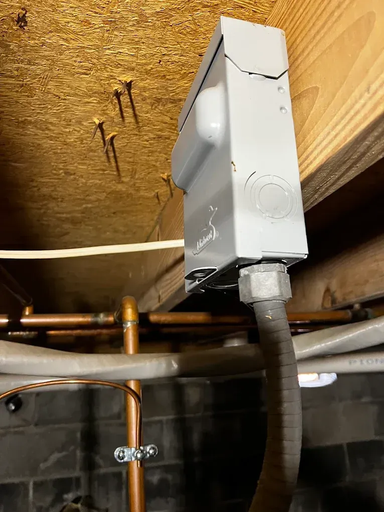 White electrical box mounted on plywood ceiling above copper pipes and ductwork in a basement.