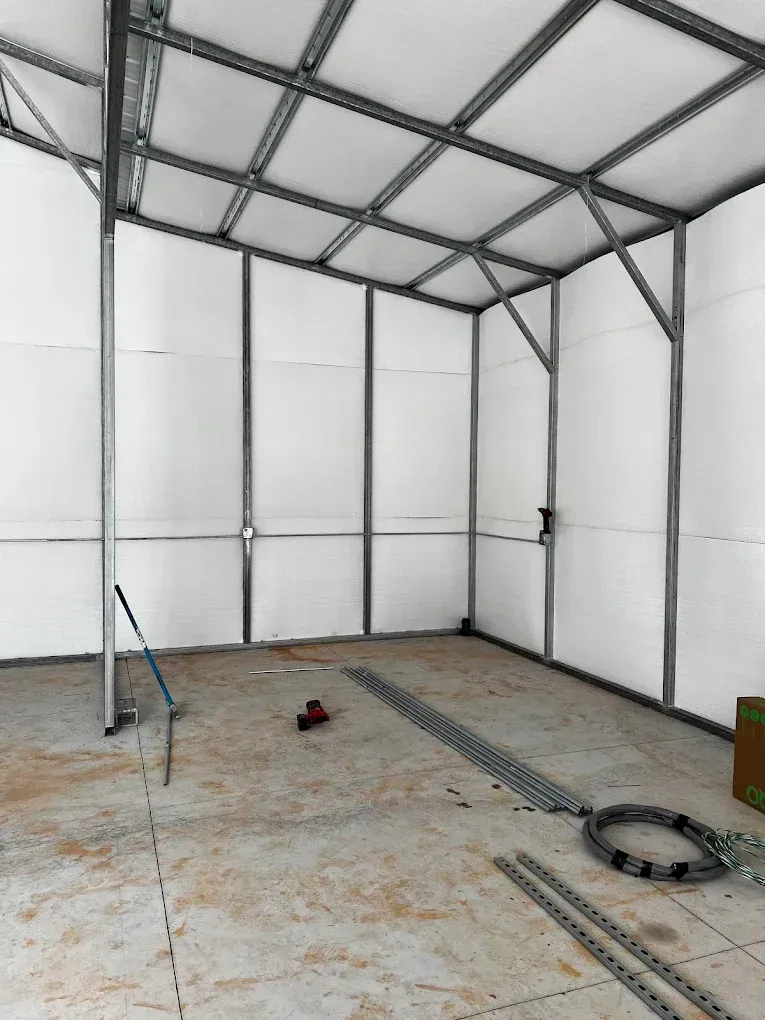 Empty metal-framed indoor tent with white wall panels and a concrete floor, scattered tools on the ground.