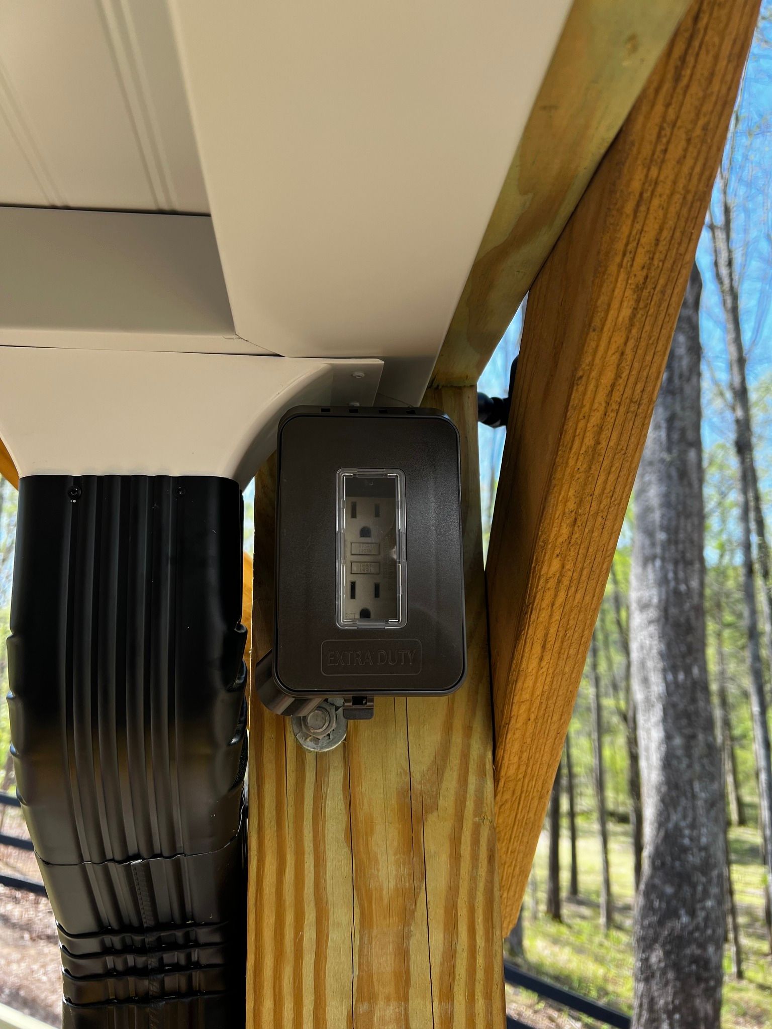 Outdoor electrical outlet mounted on a wooden post beside a screened porch with forest trees in the background