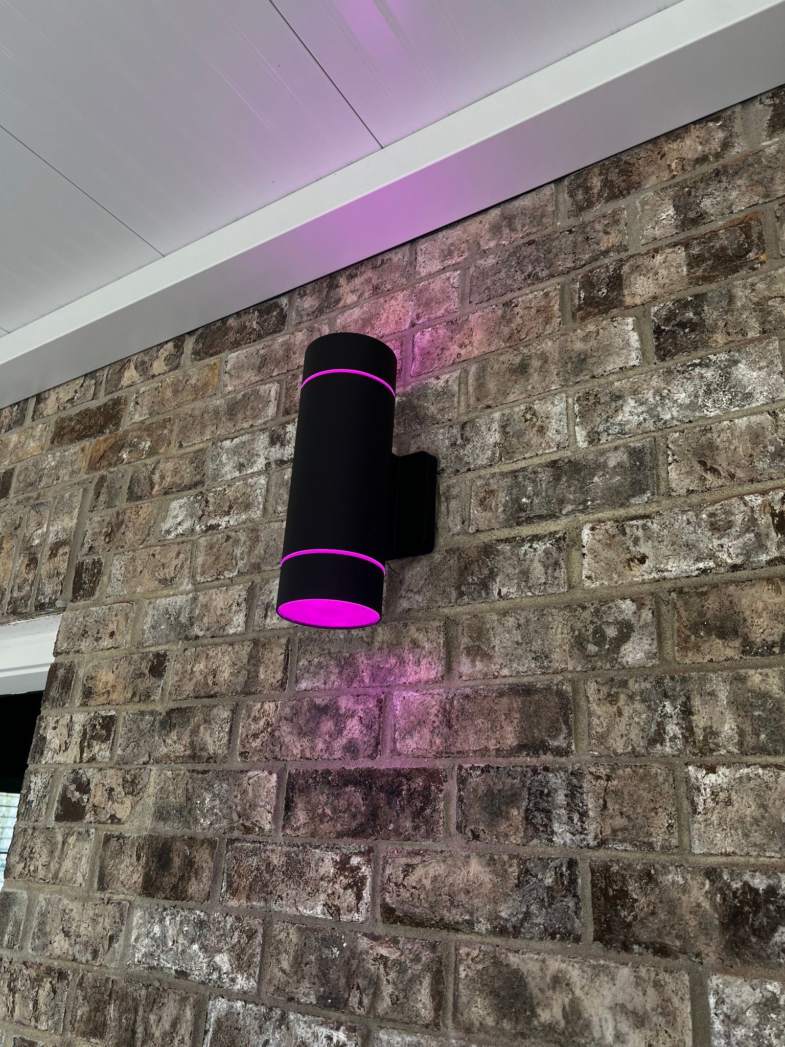 Black wall sconce with purple glow mounted on a textured brick wall, near the ceiling.