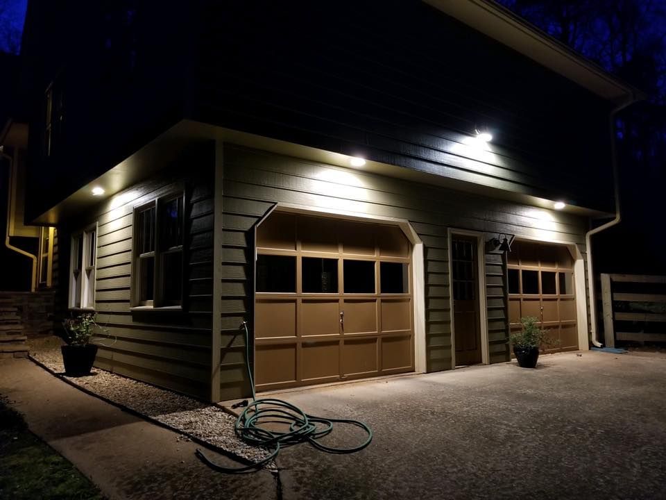 Dimly lit garage at night with closed tan doors, exterior lights, and a hose on the driveway.