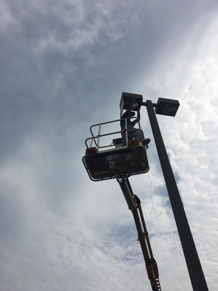 Bucket lift with a worker raised against a cloudy sky, beside a streetlight pole.