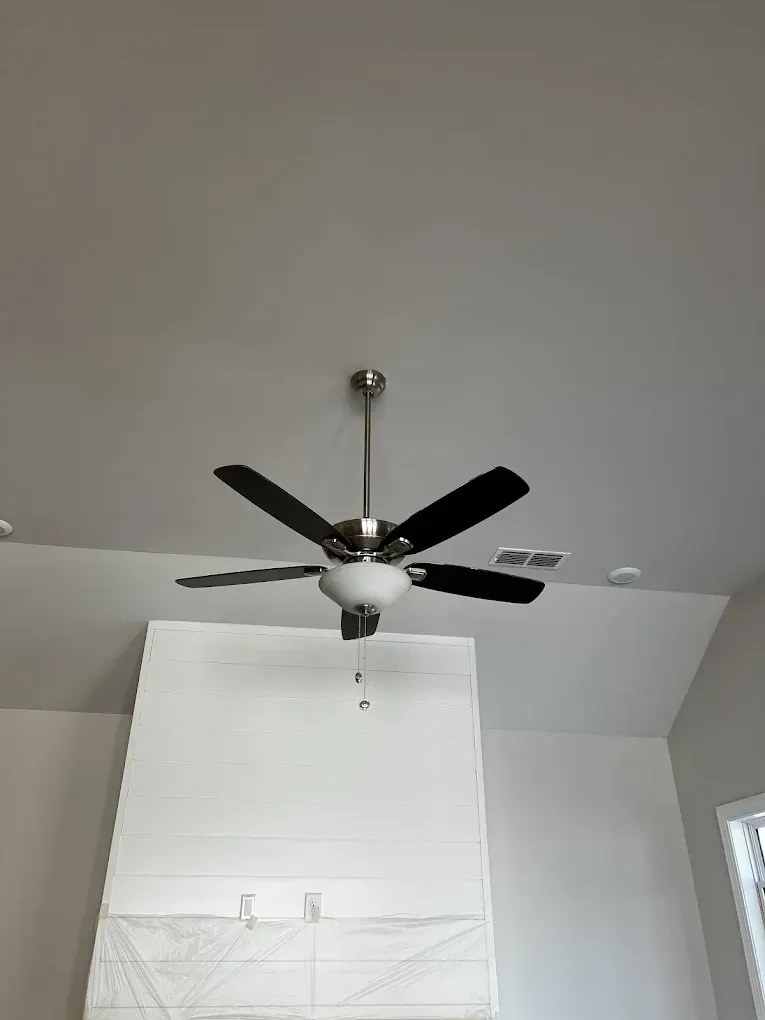 Ceiling fan with five black blades on a gray ceiling above a white-covered window.