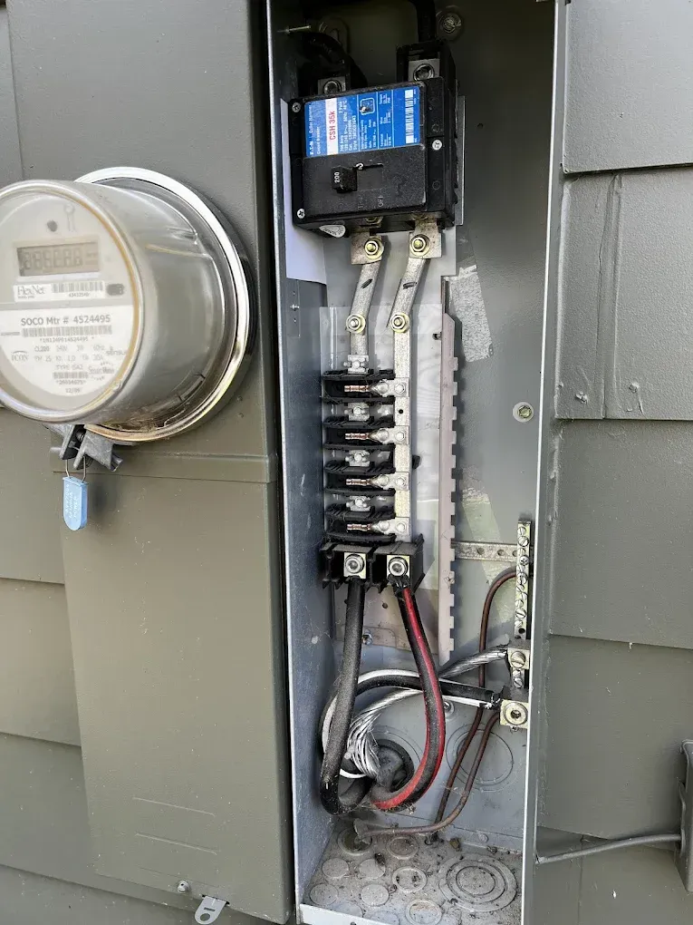 Open electrical meter panel with wiring, breakers, and a blue utility module on a gray exterior wall.