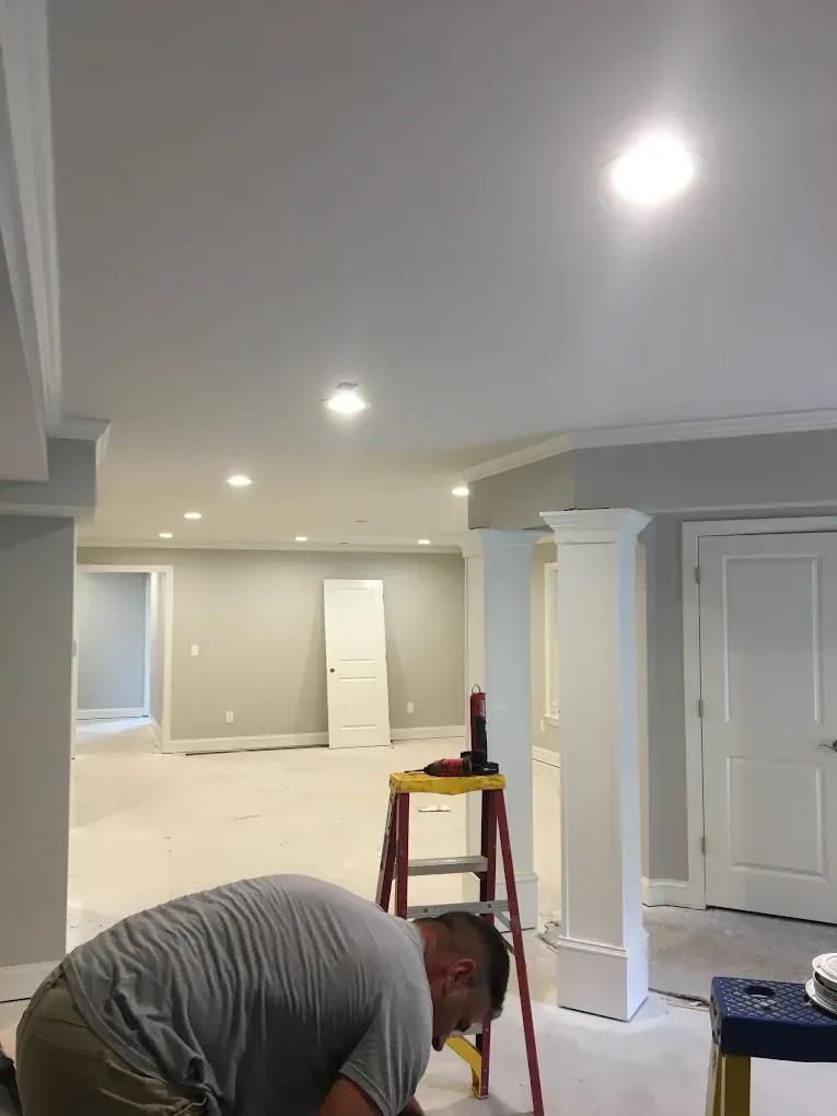 Person bending over with tools in a newly finished basement room with recessed lights and white columns