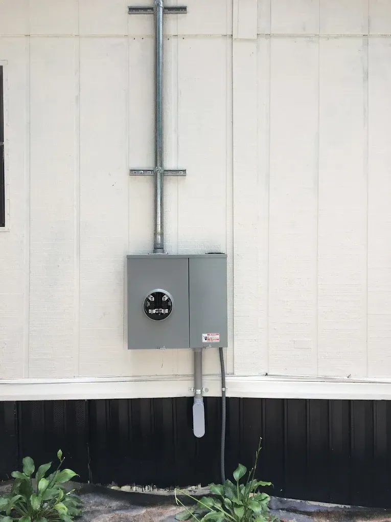 Gray electrical meter box on a white exterior wall with conduit running upward.