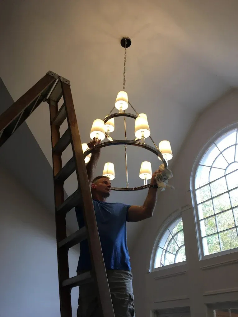 Man hanging a chandelier in a tall room beside a ladder and arched windows.