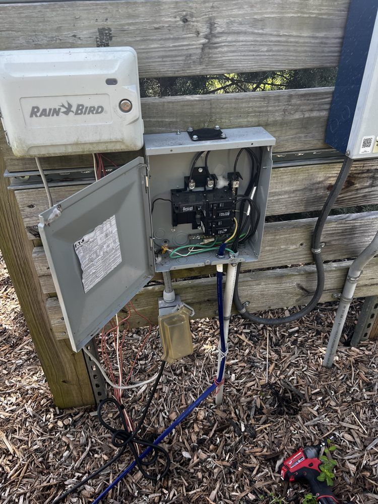 Open electrical control box beside a wooden fence, with wires and conduit exposed outdoors.