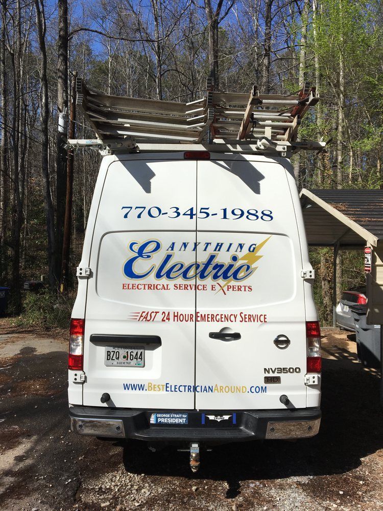 White electric company van with ladders parked on a wooded dirt road.