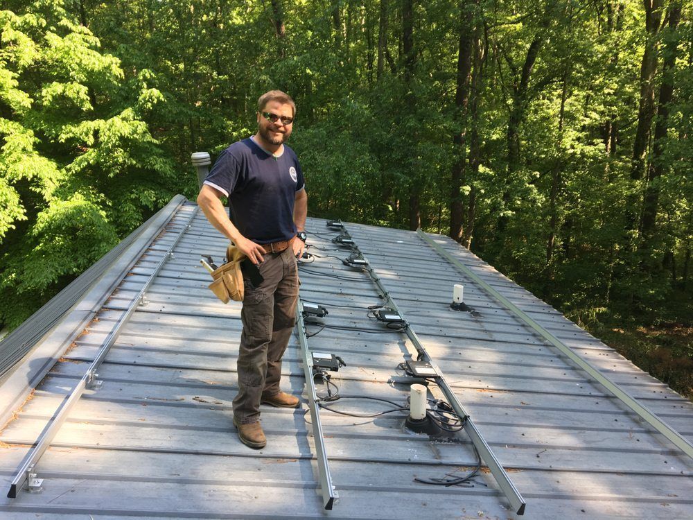 Man standing on a metal roof in a wooded area beside roof-mounted equipment and cables