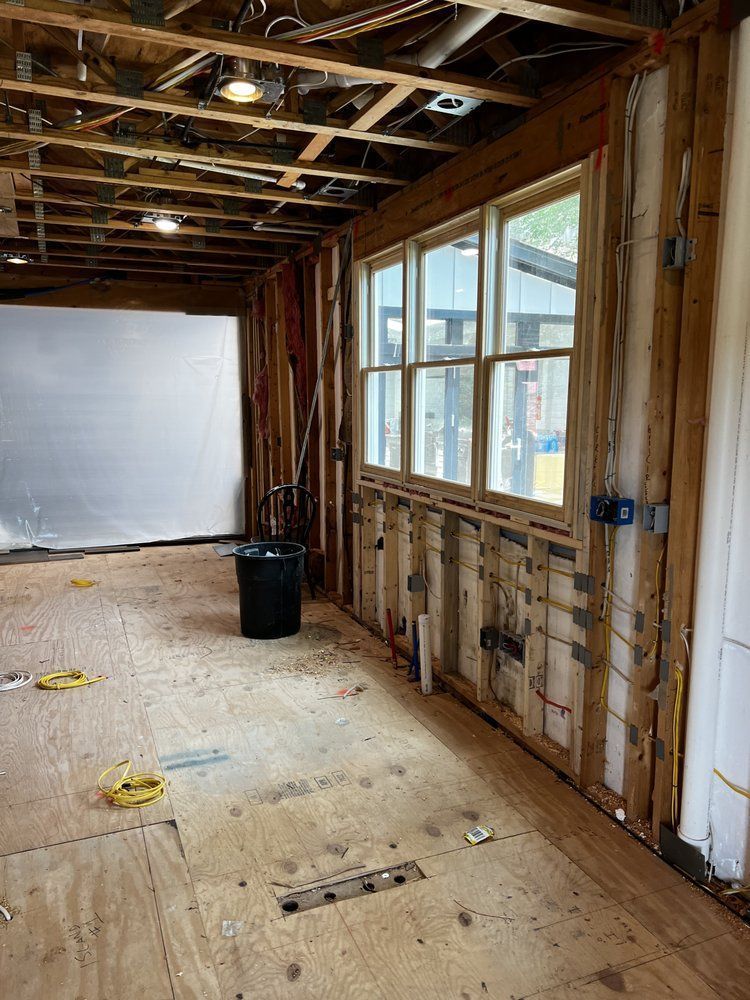 Partially renovated room with exposed framing, plywood floor, large window, and white wall covering.
