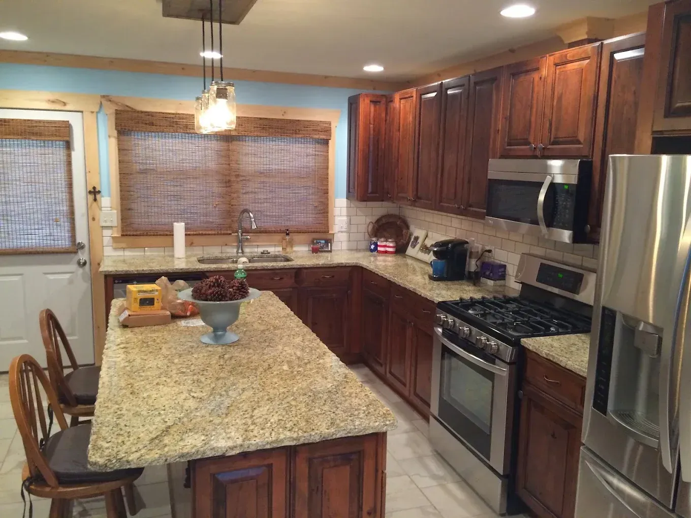 Bright kitchen with granite island, dark wood cabinets, stainless fridge, and woven blinds over the windows.