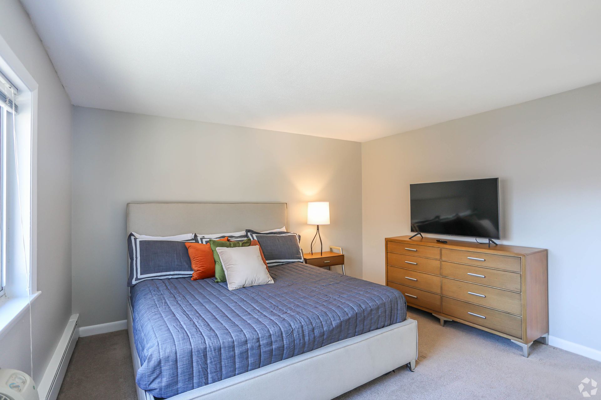 Bedroom with a bed, dresser, TV, lamp, and window, all with neutral colors.