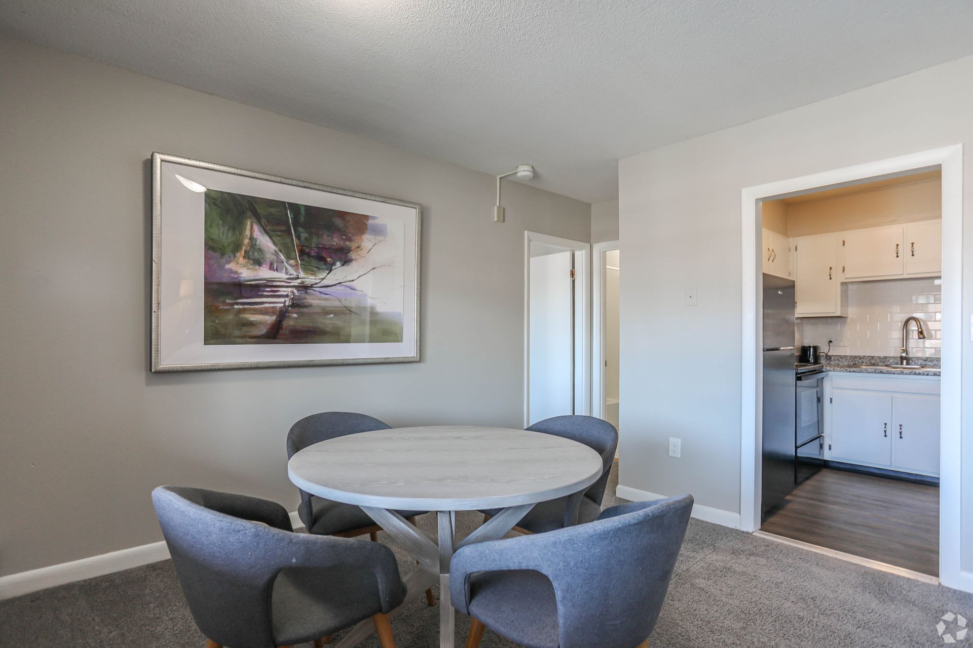 Dining area with round table, four chairs, and artwork on wall. Kitchen visible in the background.